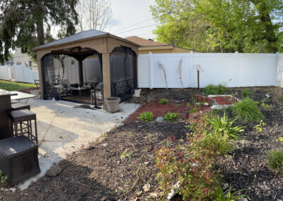 Wooden gazebo with screened sides on a concrete patio, surrounded by a white vinyl privacy fence and landscaped garden